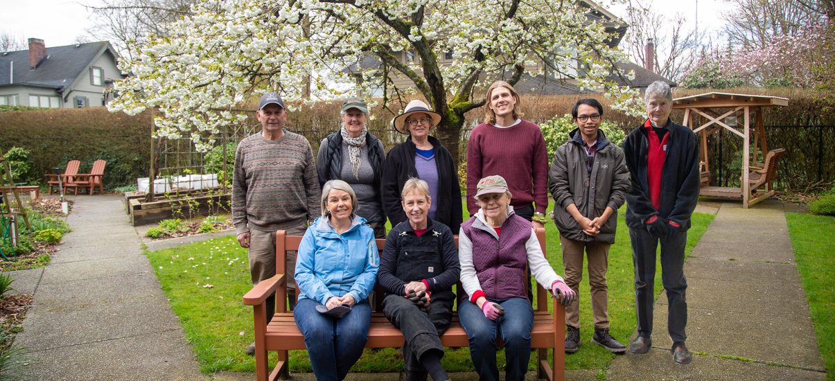 A group photo of eight Canuck Place volunteers in a lush garden setting with a blossoming tree overhead. Three individuals are seated on a wooden bench in the foreground, while five others stand behind them, smiling towards the camera. The garden has a neatly paved path, raised garden beds, wooden chairs, and a swing structure in the background.