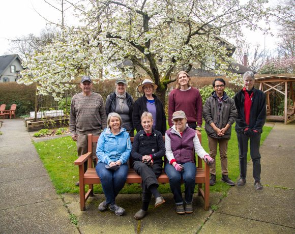 A group photo of eight Canuck Place volunteers in a lush garden setting with a blossoming tree overhead. Three individuals are seated on a wooden bench in the foreground, while five others stand behind them, smiling towards the camera. The garden has a neatly paved path, raised garden beds, wooden chairs, and a swing structure in the background.