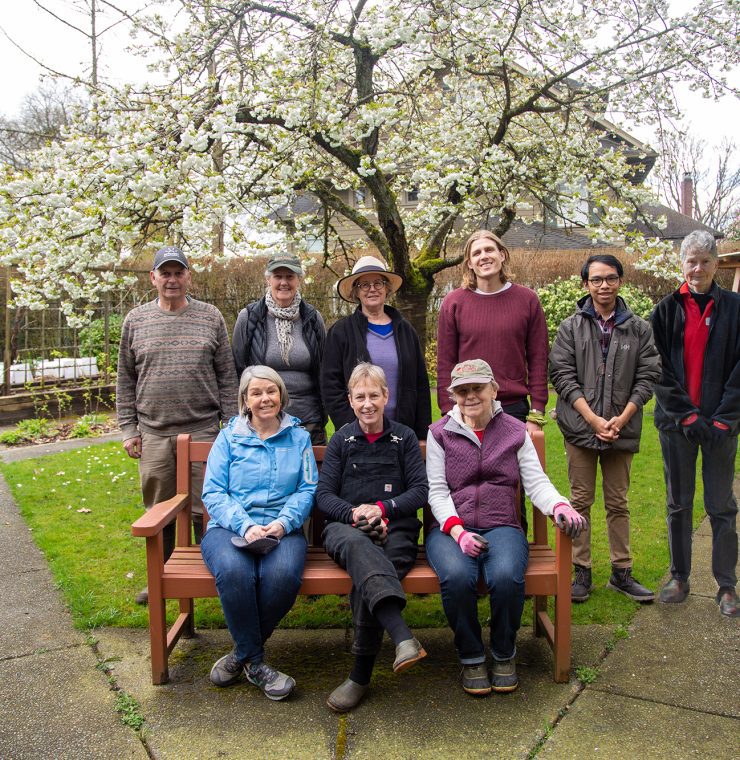 A group photo of eight Canuck Place volunteers in a lush garden setting with a blossoming tree overhead. Three individuals are seated on a wooden bench in the foreground, while five others stand behind them, smiling towards the camera. The garden has a neatly paved path, raised garden beds, wooden chairs, and a swing structure in the background.