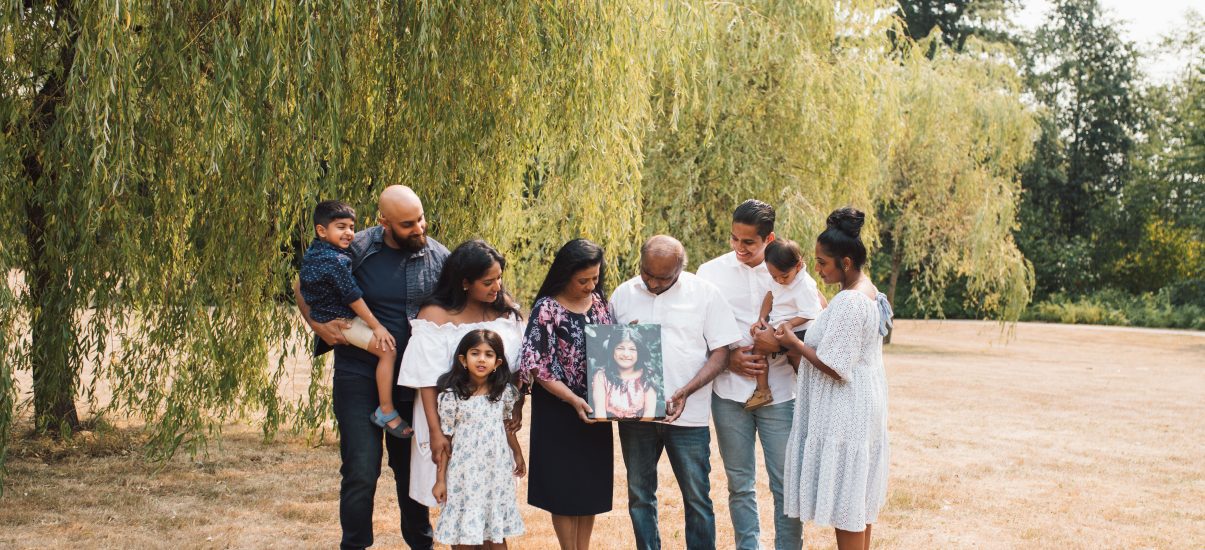 A family portrait outdoors with nine family members across three generations, posing on a grassy field with a willow tree in the background. On the left, a man with a beard holds a young boy on his shoulders. Next to him, a woman in a white off-shoulder dress smiles beside a young girl in a floral dress. In the centre, an older man and woman hold a framed photo of a teen girl, as a tribute. On the right, a woman holding a baby stands close to a man in a white shirt. They are all dressed in semi-formal attire, looking happy and connected.