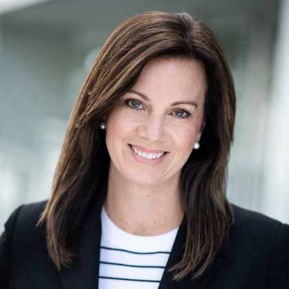A professional headshot of a woman, Canuck Place board chair Suzanne Steenburgh, with a warm and confident smile. She has long, brown hair and is wearing pearl earrings, a black blazer, and a white top with think black stripes. The background is a blurred modern office environment, suggesting a business setting.