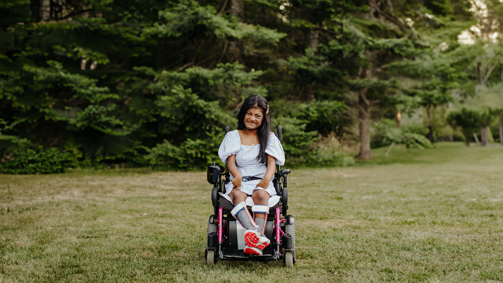 Angelina Premia, a young girl with long black hair and a warm smile sits in a wheelchair in a grassy field. She's wearing a light blue and white checkered dress with short sleeves, and leg braces. The background is softly focused, showing lush greenery and tall trees in a tranquil park setting.