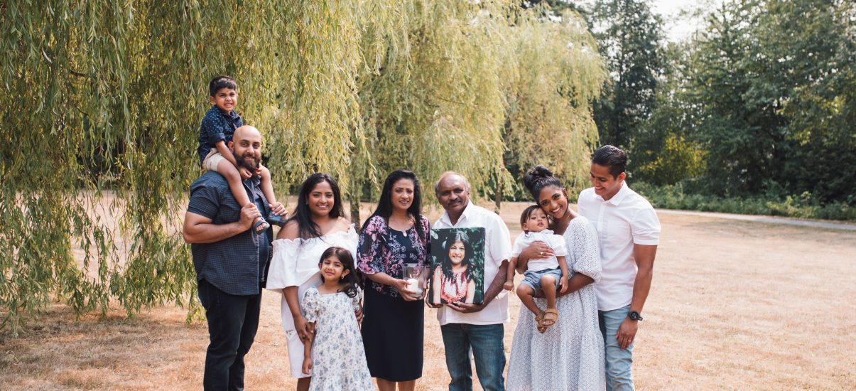 A family portrait outdoors with nine members of the Premia family across three generations, posing on a grassy field with a willow tree in the background. On the left, a man with a beard holds a young boy on his shoulders. Next to him, a woman in a white off-shoulder dress smiles beside a young girl in a floral dress. In the center, an older man and woman hold a framed photo of a teen girl, as a tribute. On the right, a woman holding a baby stands close to a man in a white shirt. They are all dressed in semi-formal attire, looking happy and connected.