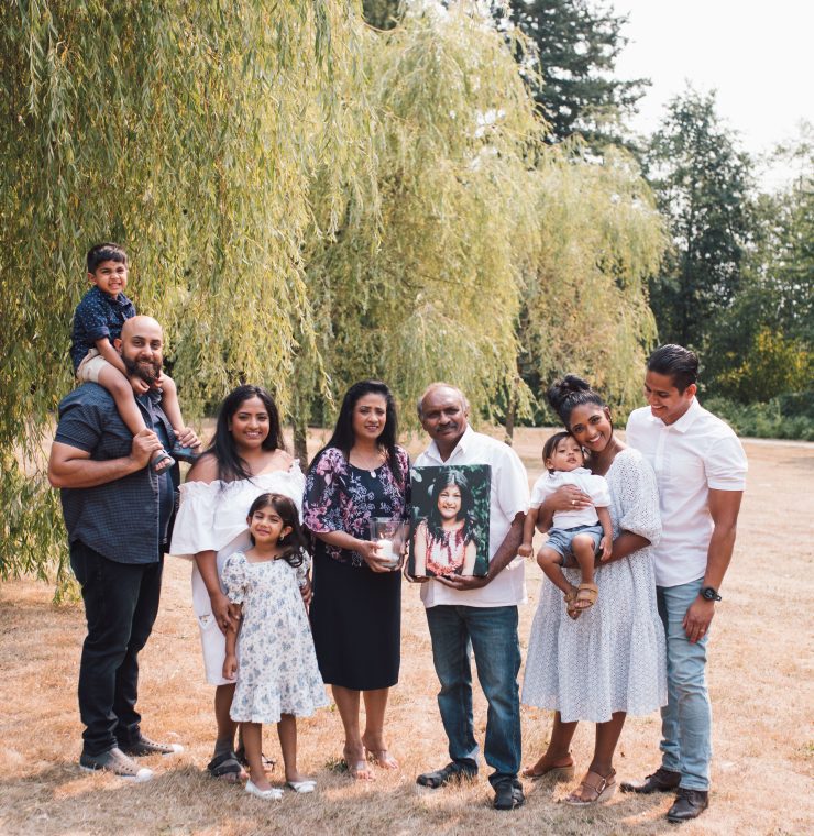 A family portrait outdoors with nine members of the Premia family across three generations, posing on a grassy field with a willow tree in the background. On the left, a man with a beard holds a young boy on his shoulders. Next to him, a woman in a white off-shoulder dress smiles beside a young girl in a floral dress. In the center, an older man and woman hold a framed photo of a teen girl, as a tribute. On the right, a woman holding a baby stands close to a man in a white shirt. They are all dressed in semi-formal attire, looking happy and connected.