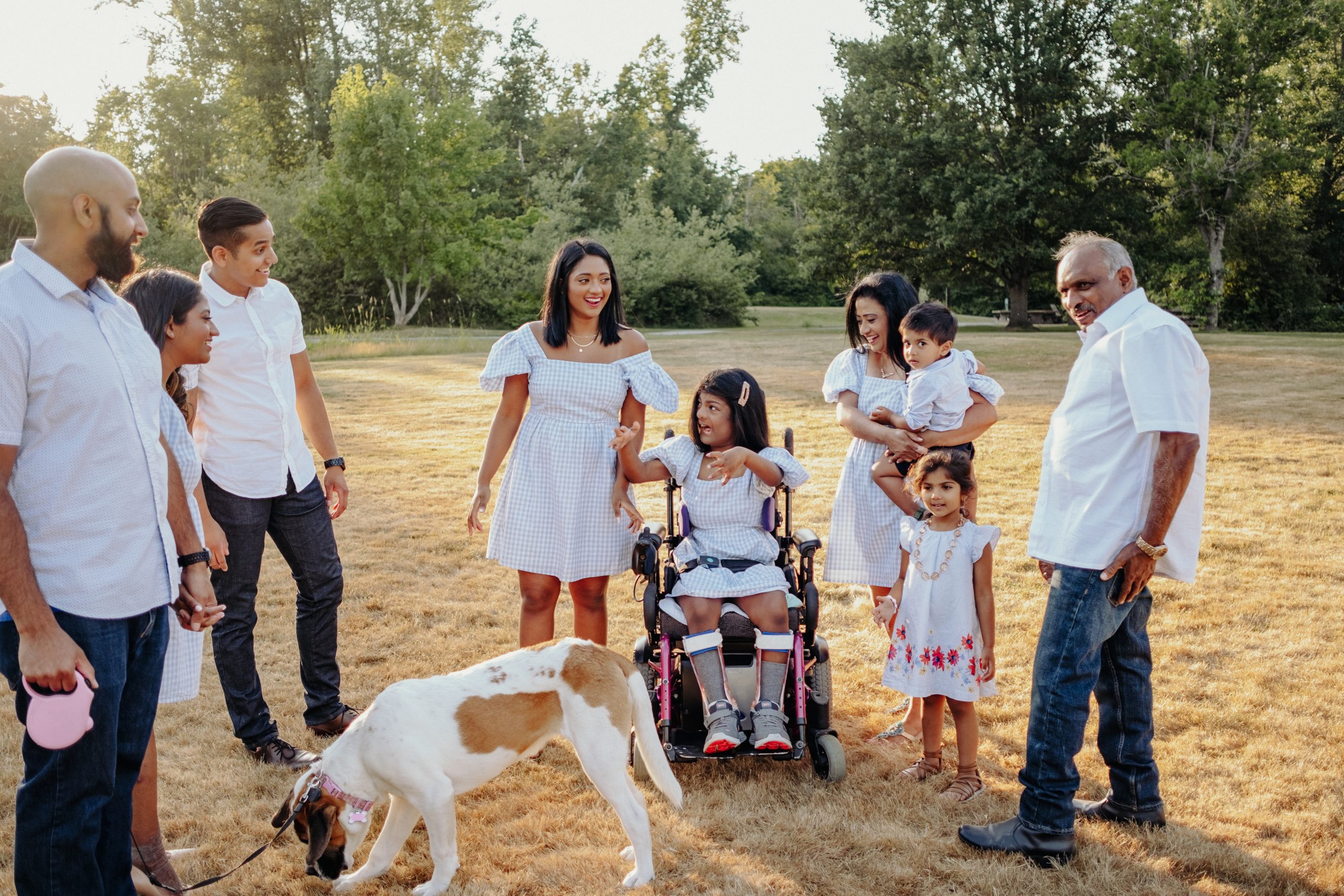 A multi-generational family is gathered in a sunlit field with trees in the background. They are dressed in coordinated outfits of white and light blue. On the left, a man holds a pink leash attached to a medium-sized brown and white dog. Beside him, a young couple holds hands, smiling towards the center where a young girl in a wheelchair is playfully interacting with a young woman in a blue dress. To the right, a woman holds a toddler, with a young girl standing in front, and an older man to the side. The mood is cheerful and familial.