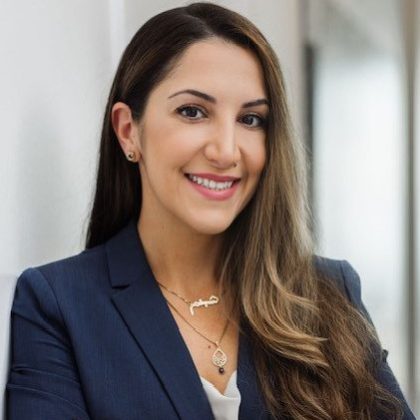 Portrait of a professional woman smiling at the camera. She has long, dark hair and is wearing a navy blue suit with a light blouse. She is wearing a silver necklace and black stud earrings. The background is a softly blurred office setting.