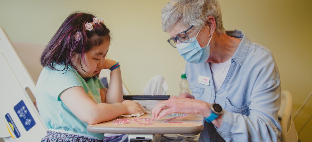 A young girl with downcast eyes is seated at a hospital bedside tray, with a Canuck Place family volunteer wearing a light blue shirt and a protective mask engaged in an activity with her. The worker is guiding the girl’s hands in a bead sorting or craft project and they both seem focused on the task at hand.