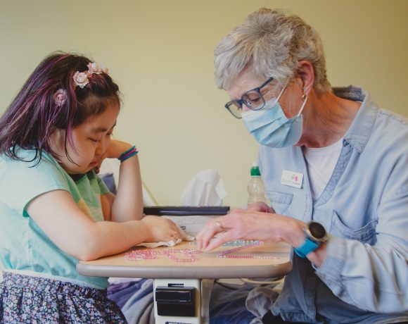 A young girl with downcast eyes is seated at a hospital bedside tray, with a Canuck Place family volunteer wearing a light blue shirt and a protective mask engaged in an activity with her. The worker is guiding the girl’s hands in a bead sorting or craft project and they both seem focused on the task at hand.