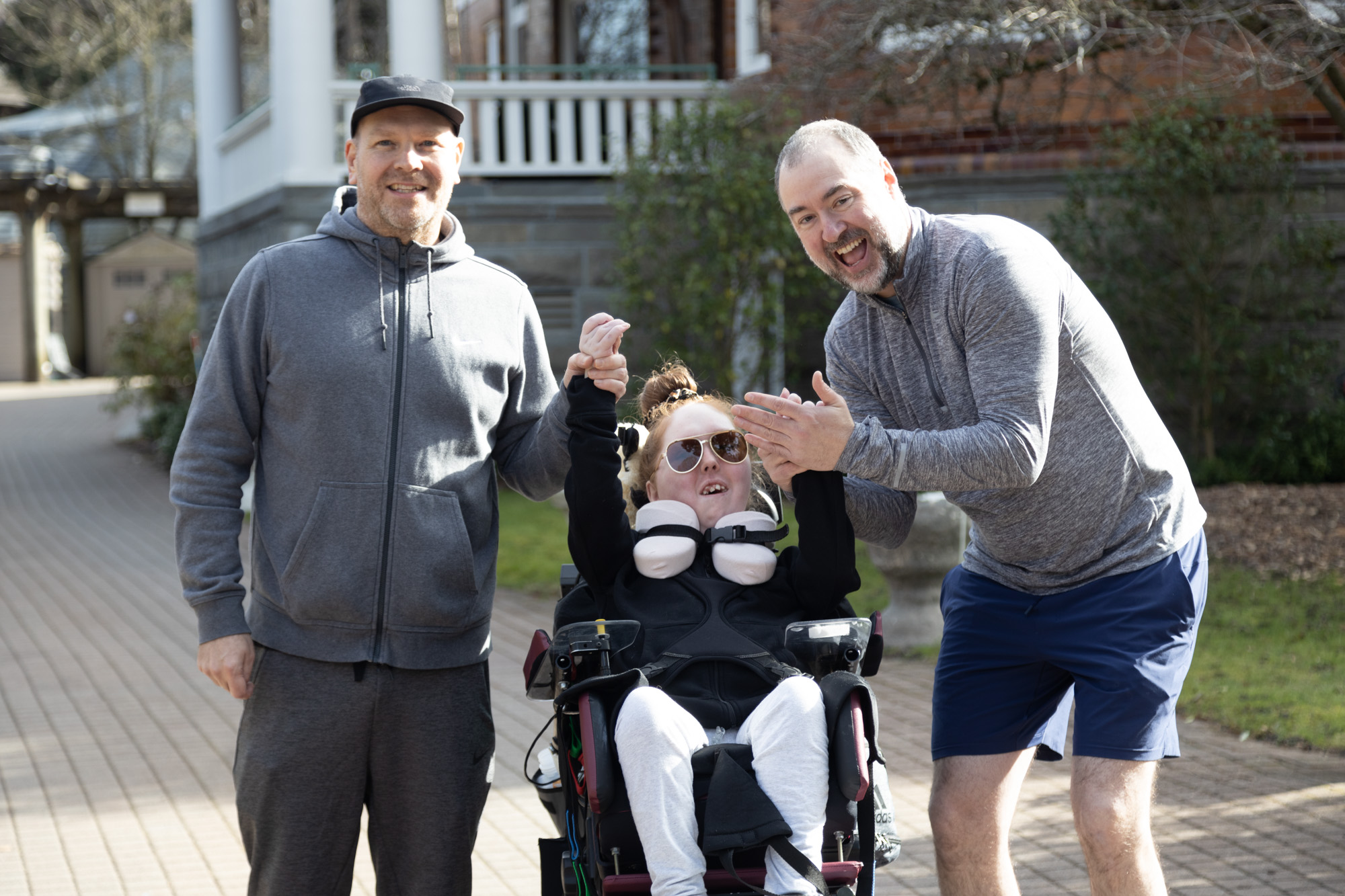 An outdoor photograph featuring three people sharing a joyful moment. On the left is a man dressed in a gray hoodie and matching sweatpants, smiling at the camera. In the center is a young girl in a wheelchair wearing sunglasses and a neck support, their arms raised mid-laughter. On the right is another man, crouching slightly, in a gray long-sleeved shirt and navy shorts, clapping and laughing along. The background is the exterior of Canuck Place Children's Hospice Vancouver - Glen Brae Manor.
