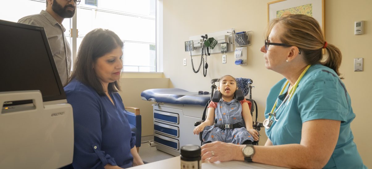 A healthcare provider sits at a table with a woman while a child in a wheelchair is sitting between them in a clinic room