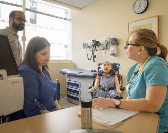A healthcare provider sits at a table with a woman while a child in a wheelchair is sitting between them in a clinic room