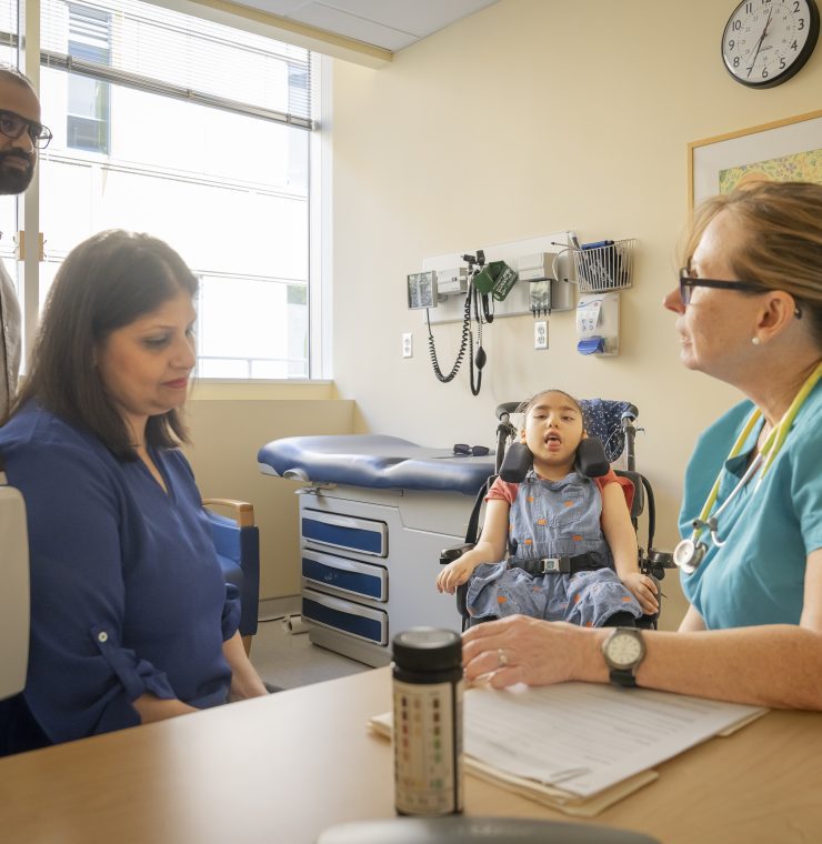 A healthcare provider sits at a table with a woman while a child in a wheelchair is sitting between them in a clinic room
