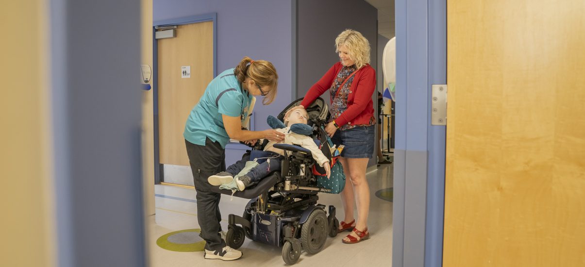 A nurse examines a child in a wheelchair in a hospital hallway while his mother stands by comforting the child