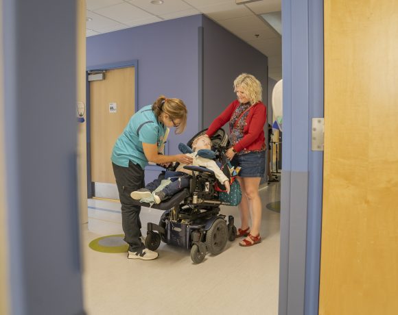 A nurse examines a child in a wheelchair in a hospital hallway while his mother stands by comforting the child