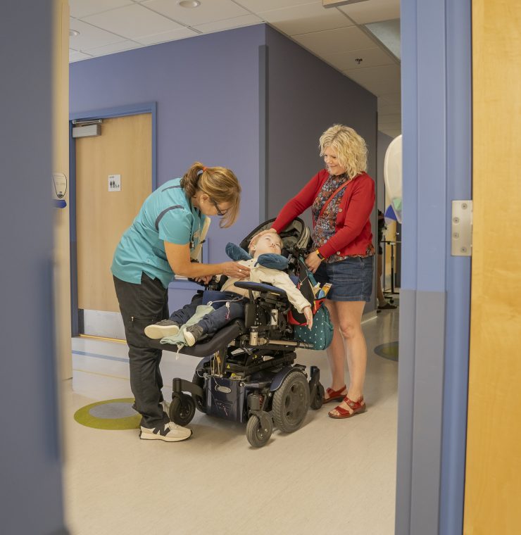 A nurse examines a child in a wheelchair in a hospital hallway while his mother stands by comforting the child