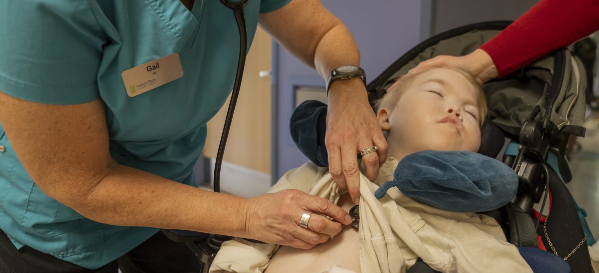 A nurse adjusts a medical device on a child in a wheelchair while the mother offers support