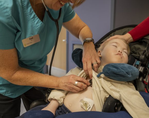 A nurse adjusts a medical device on a child in a wheelchair while the mother offers support