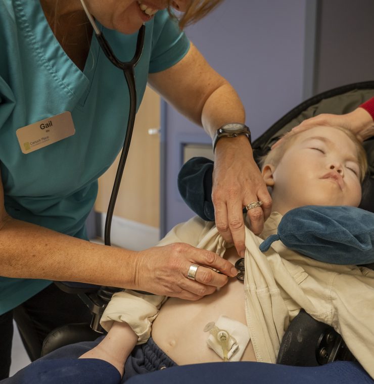A nurse adjusts a medical device on a child in a wheelchair while the mother offers support