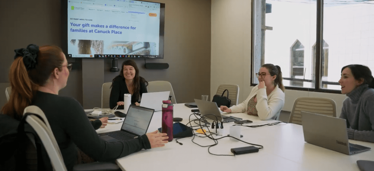 A group of four Canuck Place staff members engaged in a collaborative meeting around a conference table, filled with laptops and work materials. One woman is turned towards the camera, smiling warmly. A presentation is visible on a screen in the background. The room is well-lit with natural light streaming in from windows.