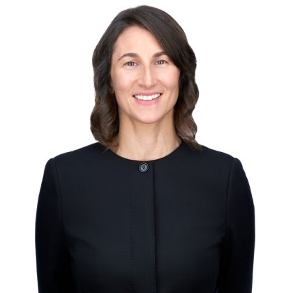 Portrait of a smiling woman with medium-length dark hair, wearing a smart black dress with a round neckline and a simple button. The background is plain white.