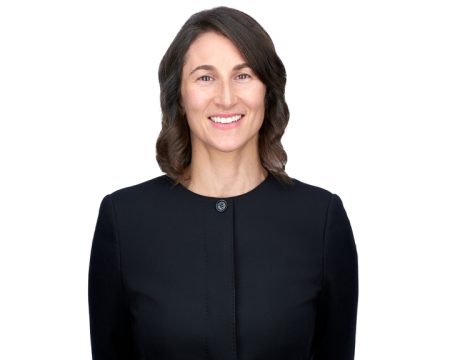 Portrait of a smiling woman with medium-length dark hair, wearing a smart black dress with a round neckline and a simple button. The background is plain white.