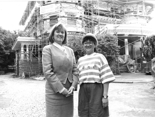 Black and white photo of two women standing side by side in front of the Canuck Place Vancouver hospice undergoing renovation, with scaffolding visible in the background. One woman is dressed in a formal business suit, and the other is in casual attire with a striped t-shirt and cap. They are both smiling at the camera.