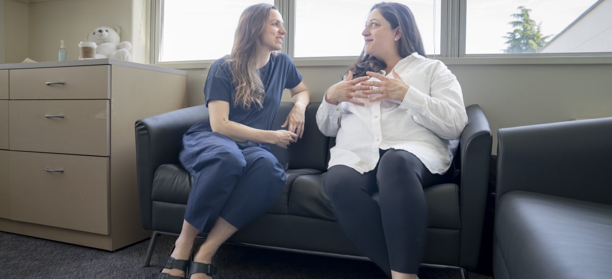 Two women sit on a couch in a meeting room gesturing while in conversation