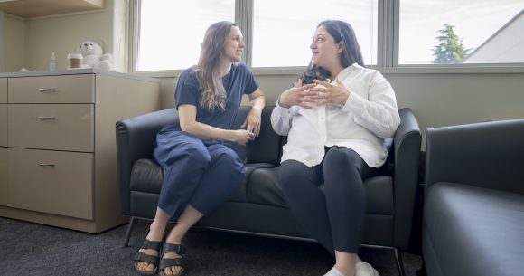 Two women sit on a couch in a meeting room gesturing while in conversation