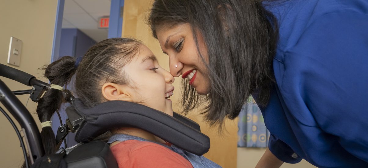 A close-up of a child in a wheelchair with their mother leaning in and smiling