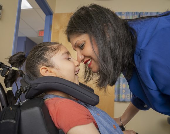 A close-up of a child in a wheelchair with their mother leaning in and smiling