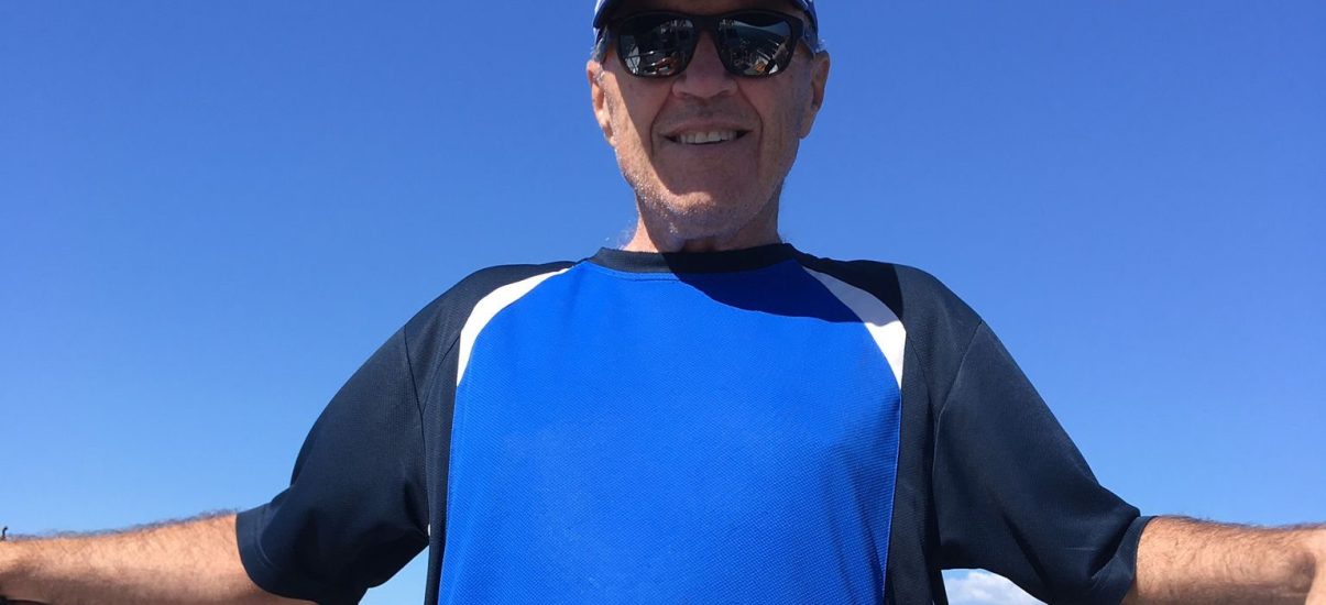 A middle-aged man in a bright blue t-shirt with dark blue sleeves and a light blue baseball cap and large black sunglasses is standing on a boat, smiling towards the camera.
