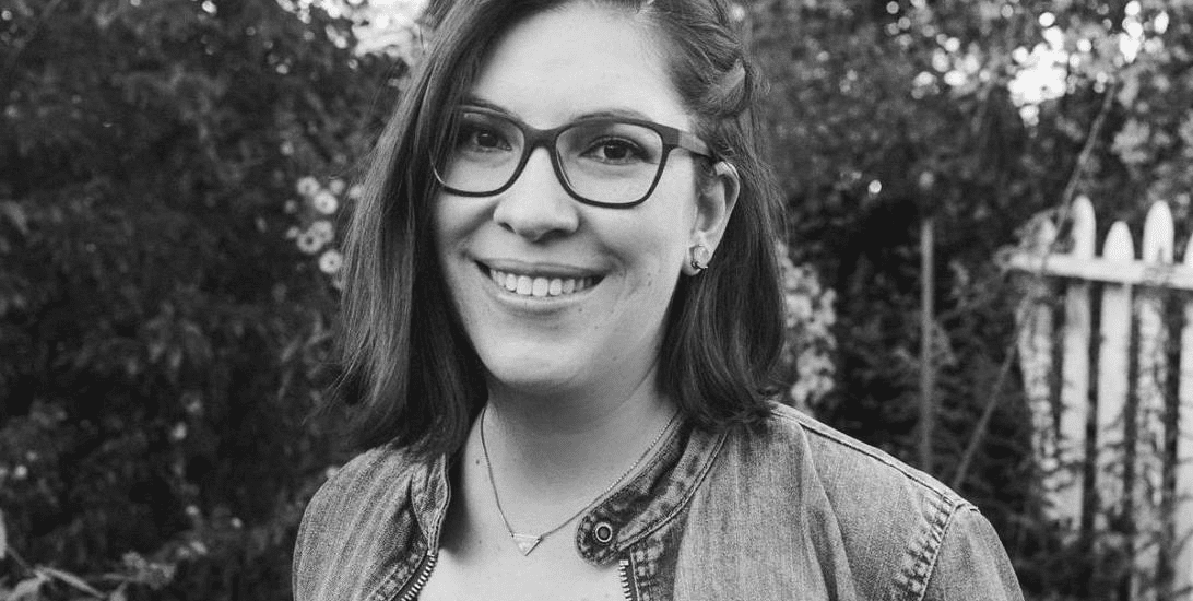 A black and white portrait of a woman smiling at the camera with greenery and a white picket fence in the background. She has shoulder-length hair and is wearing large retro square glasses, and a denim jacket over a black v-neck t-shirt.