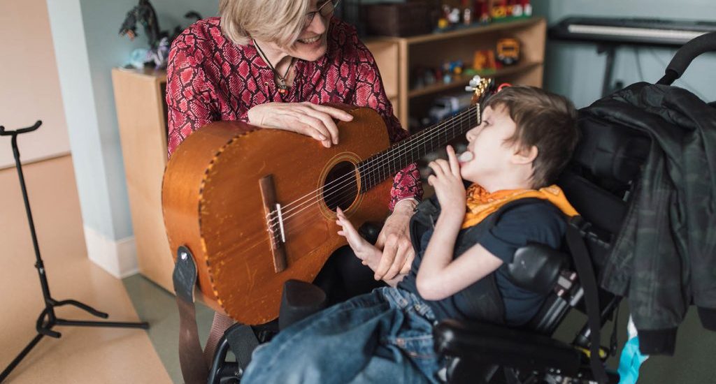 A blond-haired woman plays a guitar with a young child who is seated in a wheelchair. They are in a room filled with toys and various musical instruments.