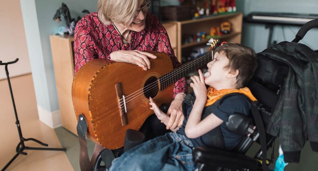 A blond-haired woman plays a guitar with a young child who is seated in a wheelchair. They are in a room filled with toys and various musical instruments.