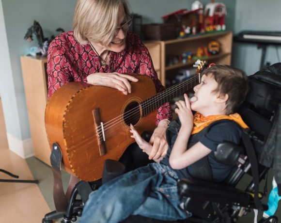 A blond-haired woman plays a guitar with a young child who is seated in a wheelchair. They are in a room filled with toys and various musical instruments.