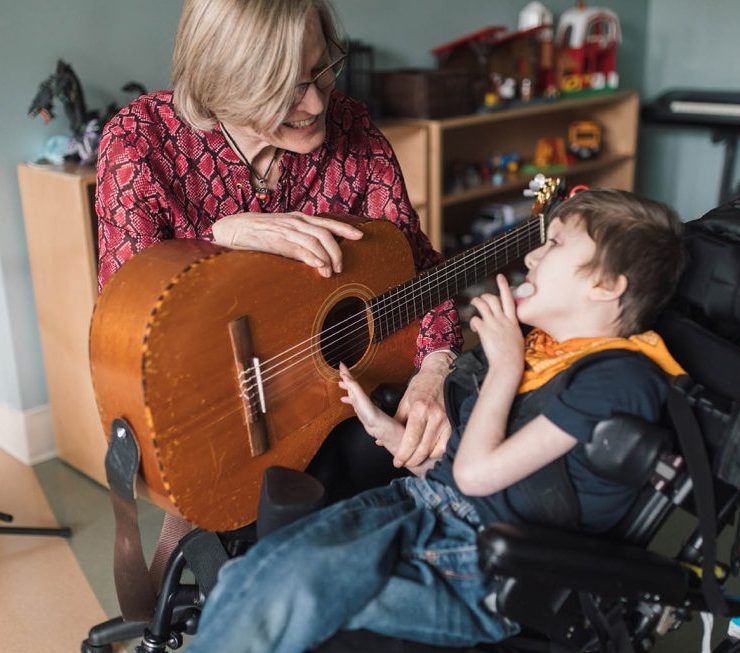 A blond-haired woman plays a guitar with a young child who is seated in a wheelchair. They are in a room filled with toys and various musical instruments.