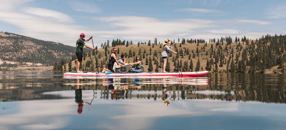 A family out on a lake on a red and white paddleboard. There is one person paddling in a standing position at each end. A child is seated at the centre of the board with one kneeling adult holding them from behind and another adult holding the child while seated in front of them.
