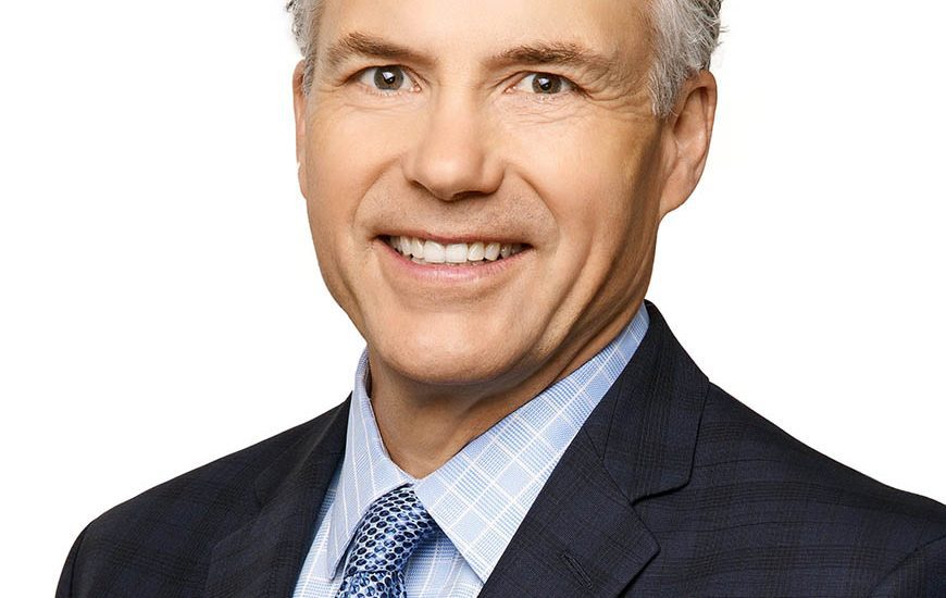 A professional headshot of a smiling middle-aged man with wavy gray hair, wearing a blue checkered shirt, blue patterned tie, and dark suit.
