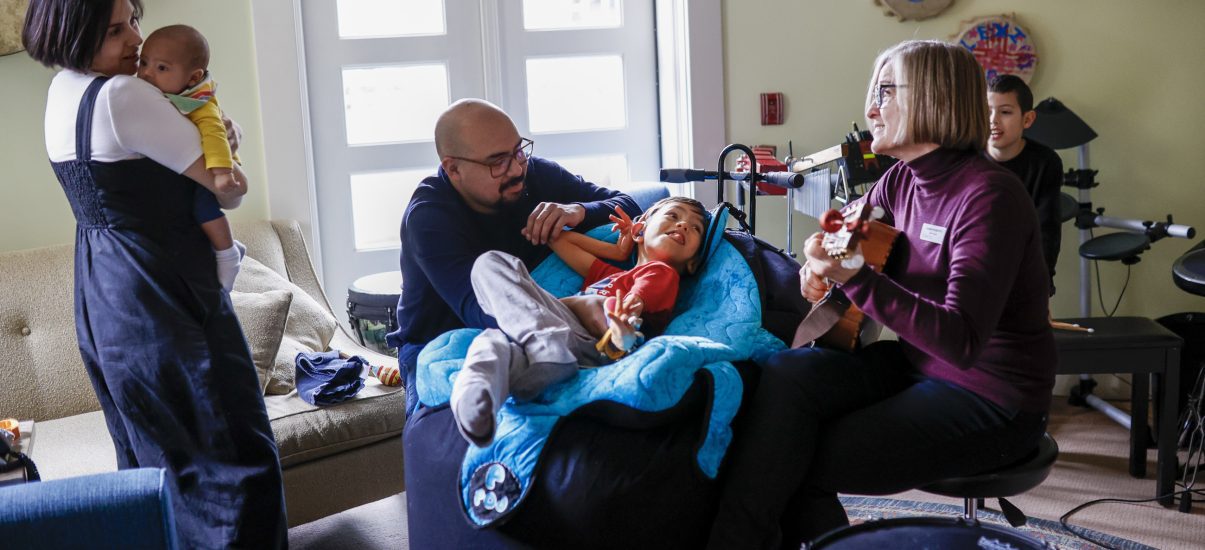 A heartwarming scene in a music therapy room at Canuck Place, where a family enjoys a musical session. A woman holding a baby stands beside a man is seated next to a boy who is laying on a bean bag. Another woman is seated to the boy’s right playing a ukulele.