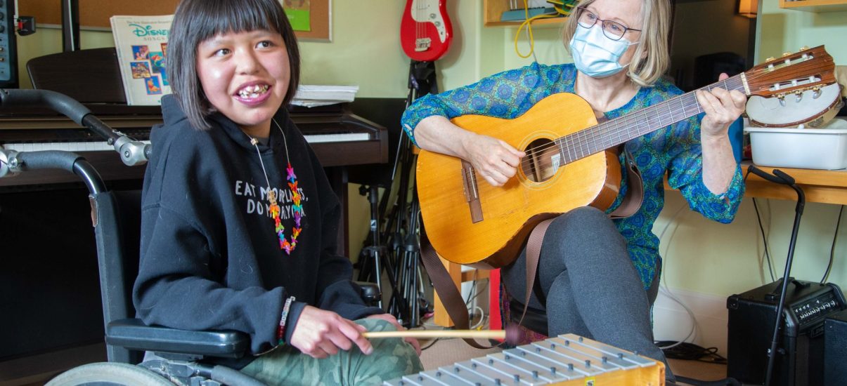 A young girl with shoulder-length dark hair and a joyful smile sits in a wheelchair, holding a small mallet and playing a wooden xylophone. She is wearing a black hoodie with colorful text and a beaded necklace. Beside her, an older white woman with short blonde hair, glasses, and a blue patterned blouse plays an acoustic guitar. She is wearing a light blue surgical mask. The setting is a music room with a piano, bookshelves filled with books and instruments, and a red electric guitar hanging on the wall. The two are engaged in a music therapy session.