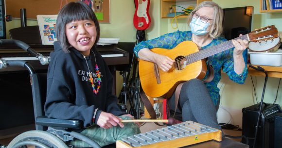 A young girl with shoulder-length dark hair and a joyful smile sits in a wheelchair, holding a small mallet and playing a wooden xylophone. She is wearing a black hoodie with colorful text and a beaded necklace. Beside her, an older white woman with short blonde hair, glasses, and a blue patterned blouse plays an acoustic guitar. She is wearing a light blue surgical mask. The setting is a music room with a piano, bookshelves filled with books and instruments, and a red electric guitar hanging on the wall. The two are engaged in a music therapy session.