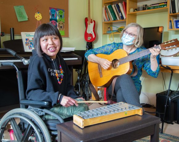 A young girl with shoulder-length dark hair and a joyful smile sits in a wheelchair, holding a small mallet and playing a wooden xylophone. She is wearing a black hoodie with colorful text and a beaded necklace. Beside her, an older white woman with short blonde hair, glasses, and a blue patterned blouse plays an acoustic guitar. She is wearing a light blue surgical mask. The setting is a music room with a piano, bookshelves filled with books and instruments, and a red electric guitar hanging on the wall. The two are engaged in a music therapy session.