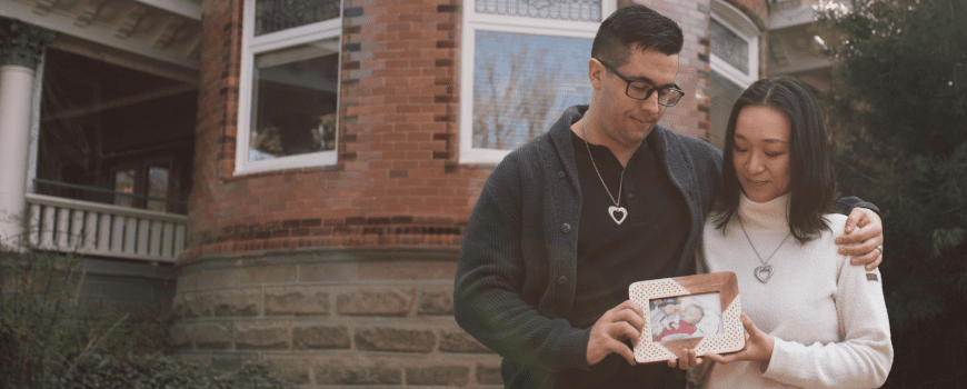 A couple stands closely together outside a brick building, each wearing a heart-shaped necklace. The man has his arm around the woman, and they both gently hold a framed photo of a baby. They look down at the picture with quiet emotion, surrounded by greenery and soft natural light.