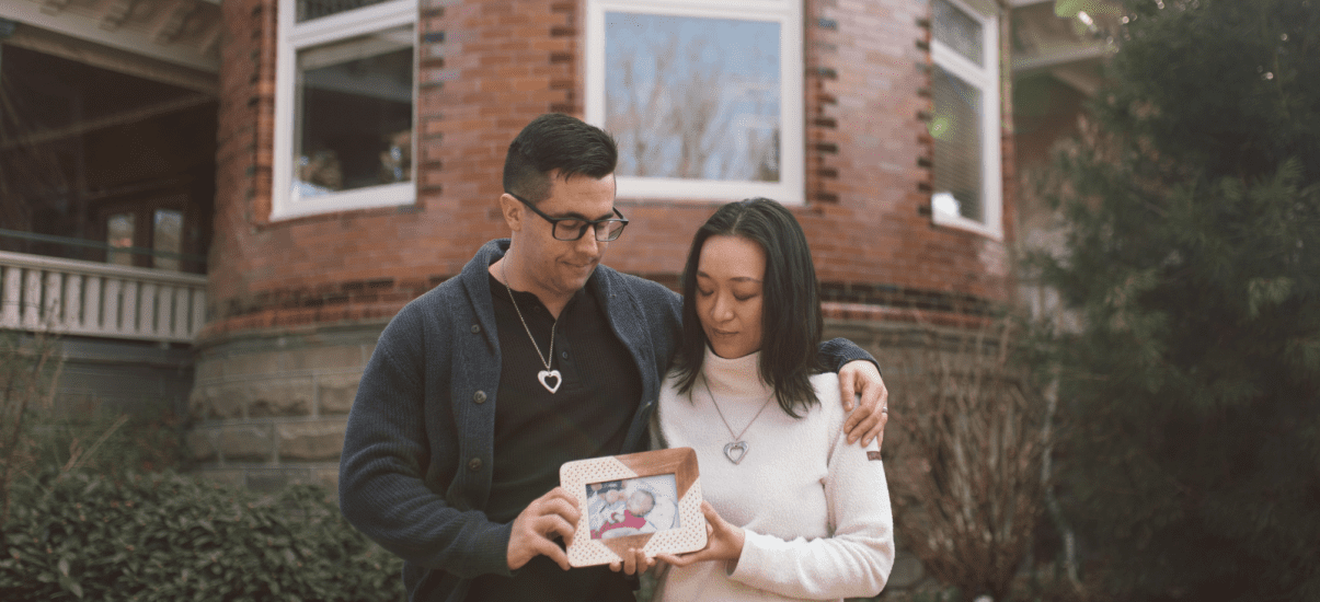 A couple stands closely together outside a brick building, each wearing a heart-shaped necklace. The man has his arm around the woman, and they both gently hold a framed photo of a baby. They look down at the picture with quiet emotion, surrounded by greenery and soft natural light.