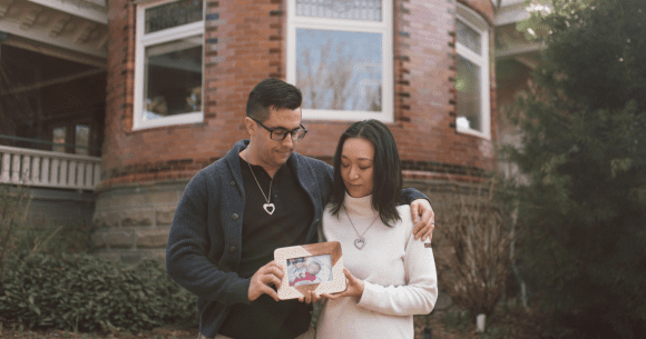 A couple stands closely together outside a brick building, each wearing a heart-shaped necklace. The man has his arm around the woman, and they both gently hold a framed photo of a baby. They look down at the picture with quiet emotion, surrounded by greenery and soft natural light.