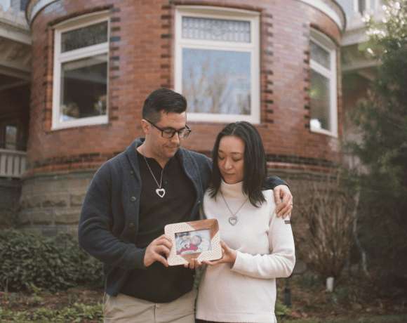 A couple stands closely together outside a brick building, each wearing a heart-shaped necklace. The man has his arm around the woman, and they both gently hold a framed photo of a baby. They look down at the picture with quiet emotion, surrounded by greenery and soft natural light.