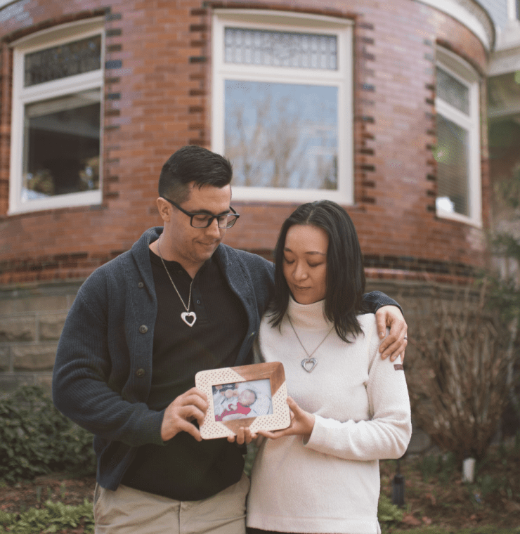 A couple stands closely together outside a brick building, each wearing a heart-shaped necklace. The man has his arm around the woman, and they both gently hold a framed photo of a baby. They look down at the picture with quiet emotion, surrounded by greenery and soft natural light.