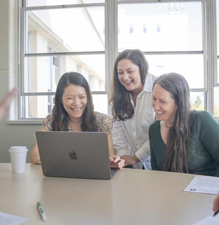 Three women gather around a laptop smiling