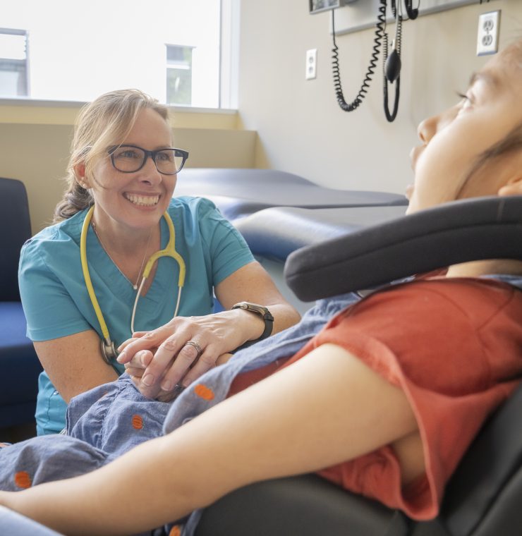 A smiling healthcare provider sits beside a child in a wheelchair in a brightly lit clinic room