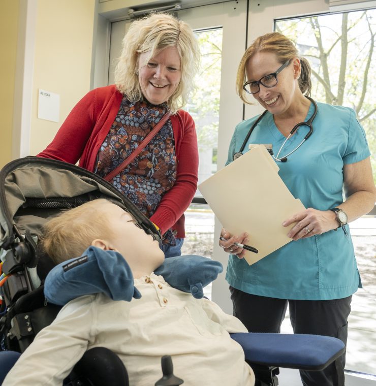 A healthcare provider interacts with a child in a wheelchair, while his mother stands beside them smiling inside a bright hospital hallway